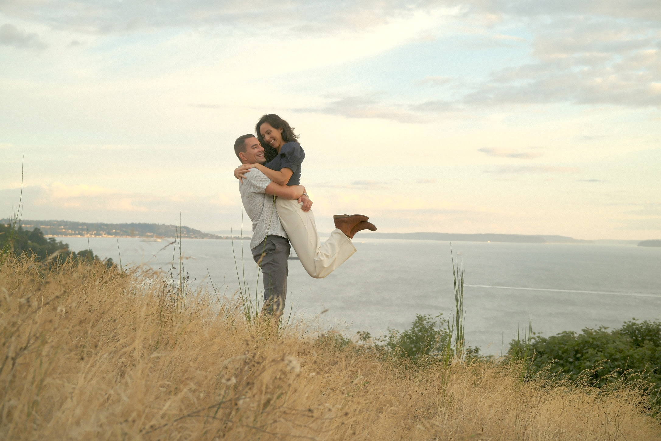 Liam lifting Caitie into a joyful embrace on a bluff overlooking the water at sunset