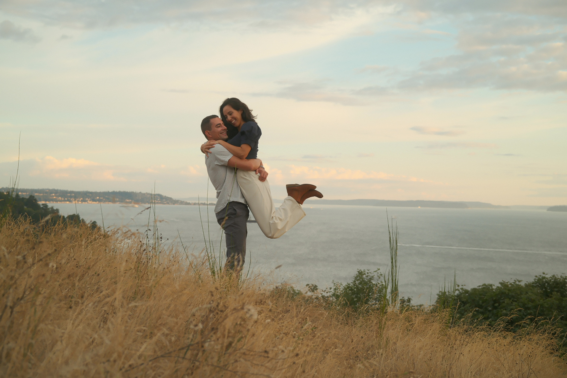 Liam lifting Caitie into a joyful embrace on a bluff overlooking the water at sunset